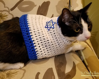 May include: A black and white cat wearing a white crochet sweater with blue trim and a Star of David emblem. The cat is sitting on a floral patterned chair. The sweater is handmade.