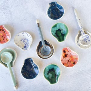 May include: A collection of handmade ceramic spoon rests and spoons in various colors and designs. The spoon rests are shell-shaped, with colors including blue, green, pink, and white. The spoons are in matching colors and patterns.