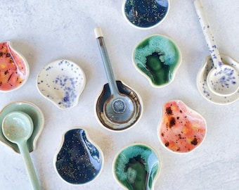 Op de afbeelding: Een verzameling handgemaakte keramische lepelsteunen en lepels in verschillende kleuren en ontwerpen. De lepelsteunen zijn schelpvormig, met kleuren als blauw, groen, roze en wit. De lepels hebben bijpassende kleuren en patronen.