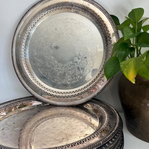 May include: A stack of silver-colored serving trays with ornate, engraved floral patterns. The trays have decorative edges and are stacked on top of each other. A green plant in a brown pot is in the background.