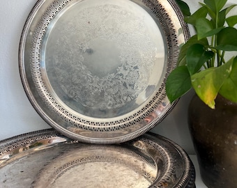 May include: A stack of silver-colored serving trays with ornate, engraved floral patterns. The trays have decorative edges and are stacked on top of each other. A green plant in a brown pot is in the background.