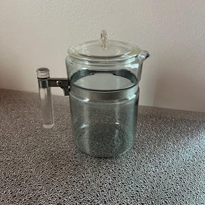 May include: A clear glass coffee pot with a lid and handle. The pot has a metal band around the middle and a clear plastic handle. The lid has a clear plastic knob. The coffee pot is sitting on a textured surface.