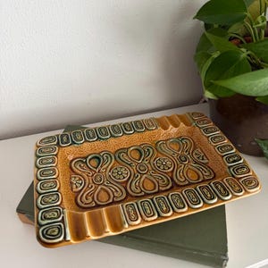 May include: A rectangular ceramic ashtray with a raised floral design in shades of brown, green, and tan. The ashtray has a textured surface and a border of oval shapes. It sits on a green book.