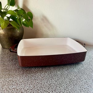 May include: A rectangular baking dish with a white interior and a dark brown exterior. The dish has two handles and is sitting on a textured surface. A potted plant is visible in the background.