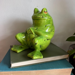 May include: A ceramic frog figurine in a relaxed pose, painted in a vibrant green with black speckles. The frog is perched atop a stack of vintage books with various colored covers, creating a whimsical decorative display.