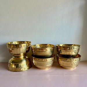May include: Six gold metal bowls of varying sizes and designs. The bowls are stacked on top of each other and are arranged in a row on a pink surface.
