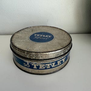 May include: Vintage Tetley Fifries tea tin. The round tin has a silver floral design on the lid and sides. The lid features a blue oval with the Tetley Fifries logo. The side of the tin has a blue band with the text "50 Tetley".