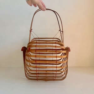 May include: A woven bamboo basket with a red handle and red string accents. The basket is empty and has a square shape.