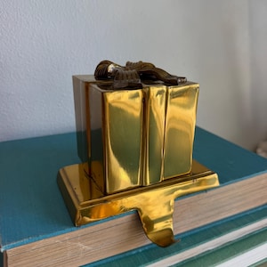 May include: A polished gold-tone metal stocking holder shaped like a square gift box. The top of the box features a sculpted bow detail, and the base includes a curved hook extending forward for hanging a stocking on a mantelpiece.