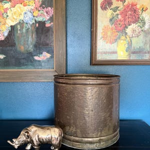 May include: A silver rhino figurine and a large, hammered metal planter sit on a dark surface. Two framed floral paintings hang on a blue wall. The planter has a wide, cylindrical shape with a textured surface.