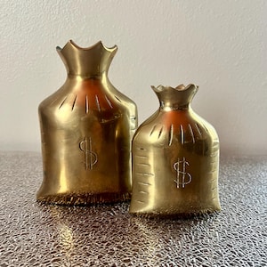 May include: Two brass money bag sculptures. The bags are gold-colored, with a dollar sign etched on the front. The bags have a ruffled top and a fringed bottom. The sculptures are displayed on a textured surface.