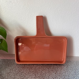 May include: A rectangular, terracotta-colored ceramic serving tray with a handle. The tray has a glossy finish and a slightly raised edge. The handle is a solid, rectangular shape, extending upwards from one end of the tray.