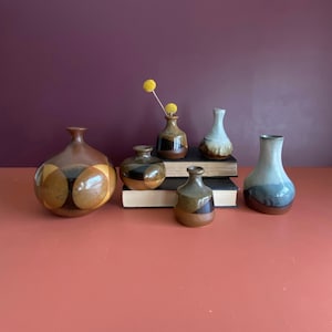 May include: A collection of six brown and black ceramic vases of varying sizes and shapes. The vases are arranged on a stack of books on a red surface.
