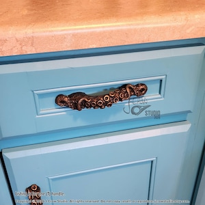 May include: A bronze-coloured cabinet handle shaped like an octopus tentacle with visible suction cups, mounted horizontally on a light blue cabinet drawer. The handle features a textured, organic design with dark highlights and a metallic finish.