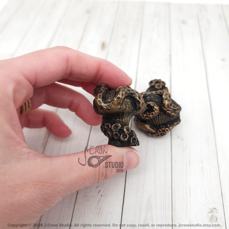 May include: Two small, dark bronze-colored cabinet knobs featuring a textured, octopus-inspired design with raised suction cup details and a weathered, metallic finish. The knobs have a rounded, organic shape with intricate, sculpted surface patterns.