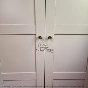 May include: A pair of white cabinet doors featuring two small, round, metallic knobs with a textured, dark bronze finish. The doors have a simple, flat-panel design with horizontal rails and vertical stiles. A watermark logo is visible in the center.