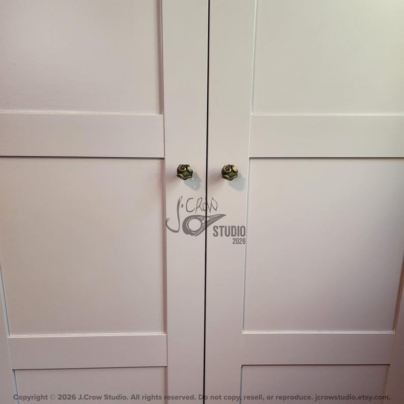 May include: A pair of white cabinet doors featuring two small, round, metallic knobs with a textured, dark bronze finish. The doors have a simple, flat-panel design with horizontal rails and vertical stiles. A watermark logo is visible in the center.