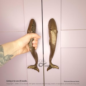 May include: A pair of bronze-finished metal cabinet handles shaped like whales, featuring detailed textures on the head and a smooth, elongated body tapering to a tail fin. The handles are mounted vertically on a light purple cabinet door surface.