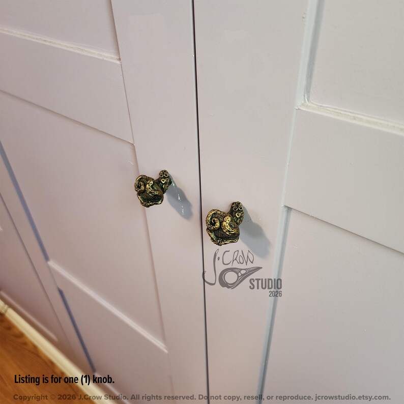 May include: Two antique brass-colored cabinet knobs shaped like coiled octopus tentacles attached to white cabinet doors. The knobs feature a textured, dark-toned finish with visible suction cup details. A logo for J. Crow Studio 2026 is visible.
