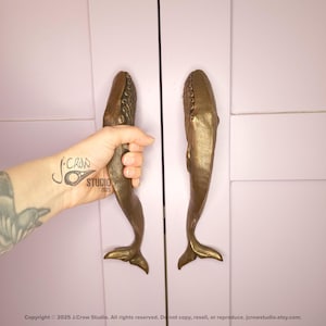 May include: Bronze whale-shaped cabinet pulls mounted on a light pink cabinet door. The pulls feature detailed fins and textured bodies. A hand is holding one of the pulls. The image includes the studio copyright. The pulls are approximately 20 cm in length.
