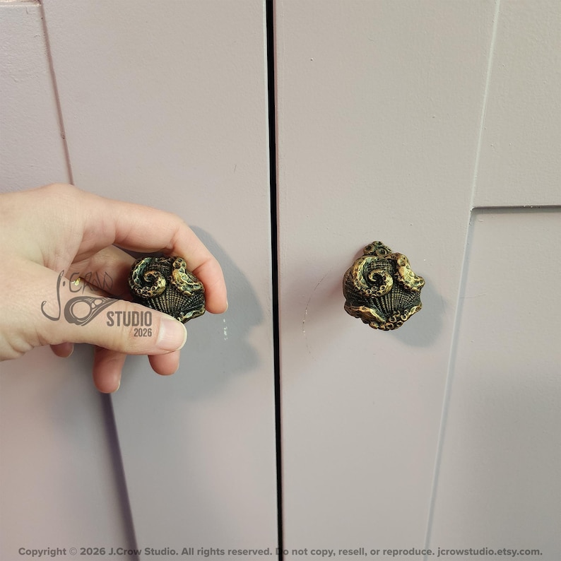 May include: A round cabinet knob with a textured, antique brass finish featuring a detailed octopus tentacle design wrapped around a shell-like base. The knob is mounted on a light lavender cabinet door, with a matching knob held nearby.