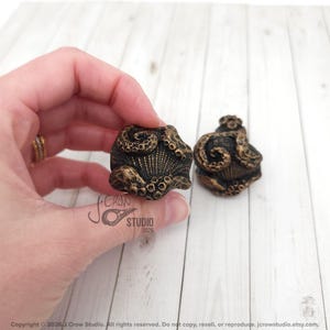 May include: Two cabinet knobs featuring a sculpted octopus design with textured tentacles wrapping around a central shell motif. The knobs have a dark, antiqued bronze finish with highlighted gold-toned details on the raised surfaces.