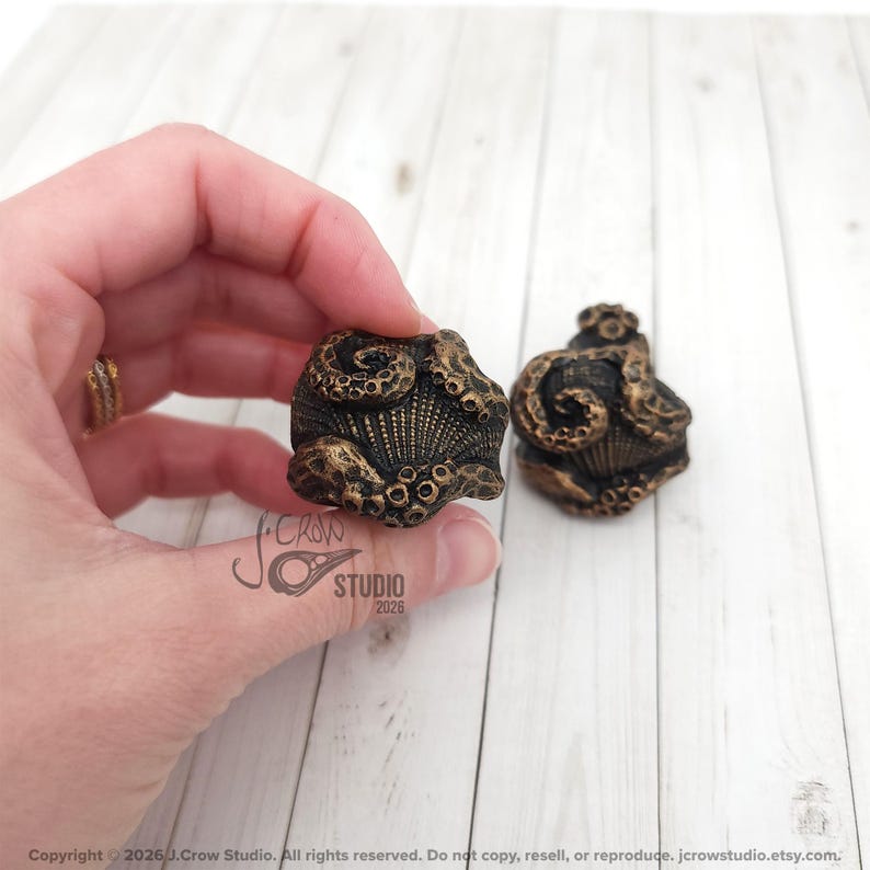May include: Two cabinet knobs featuring a sculpted octopus design with textured tentacles wrapping around a central shell motif. The knobs have a dark, antiqued bronze finish with highlighted gold-toned details on the raised surfaces.