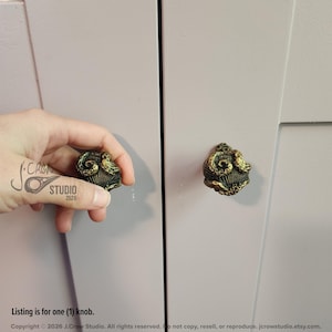 May include: A round cabinet knob featuring a detailed, three-dimensional brass-toned octopus design with textured tentacles wrapped around a shell-like base. The knob is mounted on a light lavender-colored cabinet door surface.