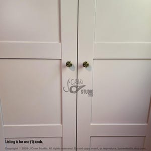 May include: Two small, round, metallic gold-toned cabinet knobs with a textured, swirling surface design attached to a pair of white, paneled cabinet doors. The text J.CROW STUDIO 2026 is visible in the center, and the text Listing is for one (1) knob.