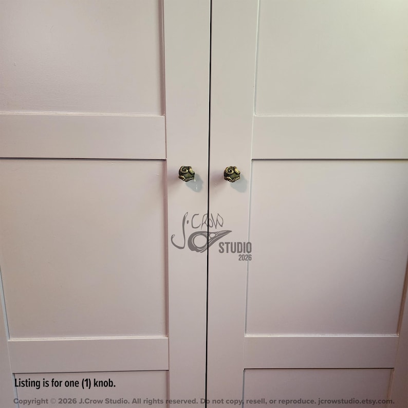 May include: Two small, round, metallic gold-toned cabinet knobs with a textured, swirling surface design attached to a pair of white, paneled cabinet doors. The text J.CROW STUDIO 2026 is visible in the center, and the text Listing is for one (1) knob.