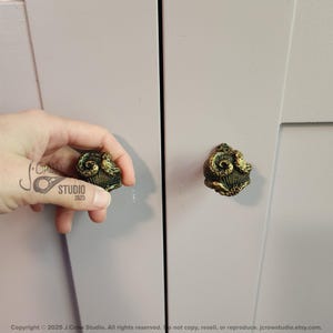 May include: Two antique gold cabinet knobs with a detailed design of an octopus or squid. The knobs are mounted on a light purple cabinet door.