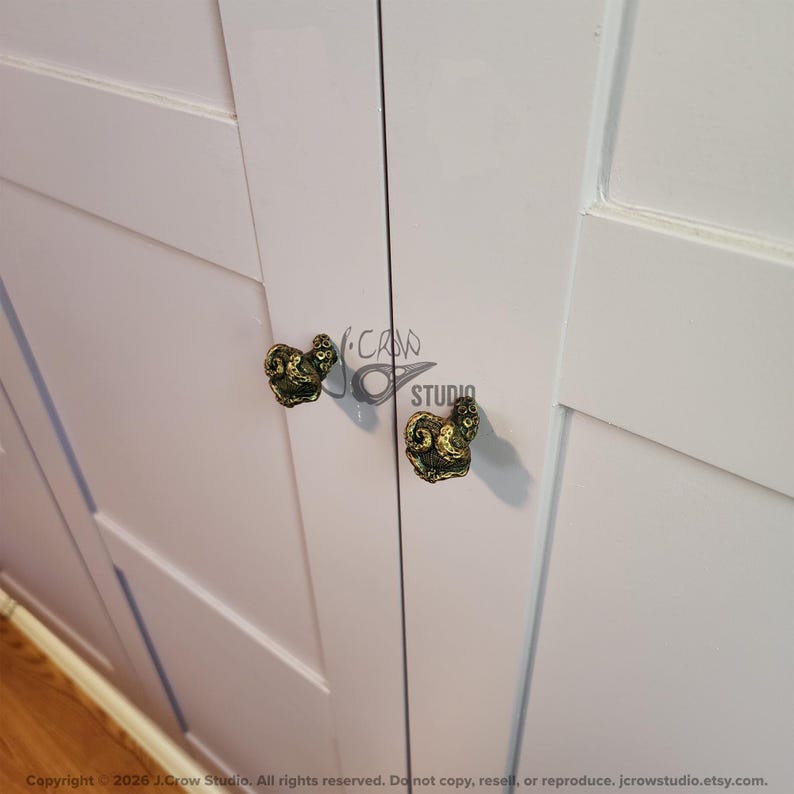 May include: Two antique brass-colored cabinet knobs shaped like curled octopus tentacles attached to a light gray cabinet door. The knobs feature detailed suction cup textures and a dark, weathered finish. A logo with the text J.CROW STUDIO is visible.