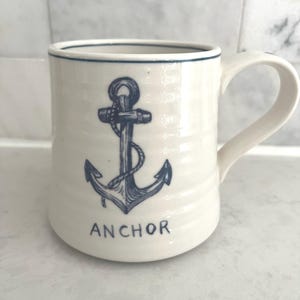 May include: Cream-colored ceramic mug with a navy blue anchor design and the word "ANCHOR" below it. The mug has a curved handle and a navy blue rim. The anchor is detailed with rope and shading.