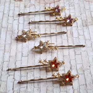 May include: Six gold-toned bobby pins with star-shaped embellishments. Each pin features a central star with surrounding smaller stars and decorative accents. The stars are adorned with clear, purple, and red crystals.