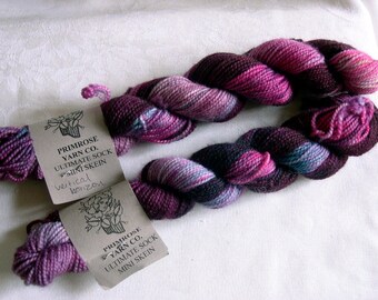 May include: Two twisted mini skeins of Primrose Yarn Co. Ultimate Sock yarn featuring a variegated color palette of deep purple, magenta, and hints of blue. Each skein has a paper label with the brand name and product text.