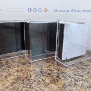May include: Three clear acrylic display cases with black, gray, and white backgrounds. The cases are empty and ready to be filled with miniature pop culture scenes.