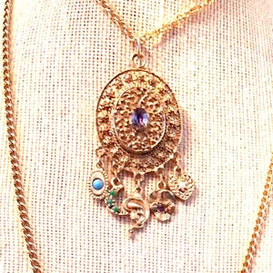 May include: Gold-toned pendant necklace with an oval filigree design, featuring a central purple gemstone and dangling charms. Charms include a turquoise stone, green accents, and a small pearl. The necklace hangs on a delicate gold chain.