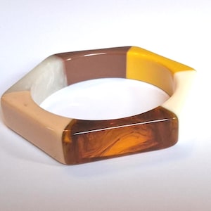 May include: A hexagonal bangle bracelet featuring a color-blocked design with segments of solid beige, translucent amber, opaque brown, bright yellow, and a marbled off-white resin material, creating a smooth, polished, and multi-toned appearance.