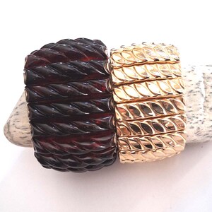 May include: Two wide stretch bracelets featuring a repeating diagonal rope-like texture. One bracelet is a deep, translucent dark brown, while the other is a polished, reflective metallic gold tone. Both items have a segmented, flexible design.