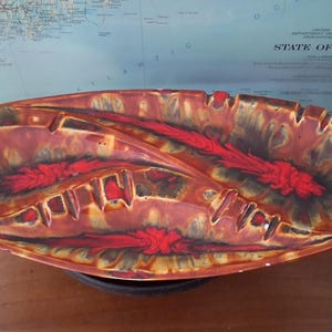 May include: An elongated oval ceramic dish featuring a vibrant glaze with swirling patterns of deep red, burnt orange, and dark brown. The surface has a glossy finish with raised ridges and indentations along the rim, creating a textured appearance.