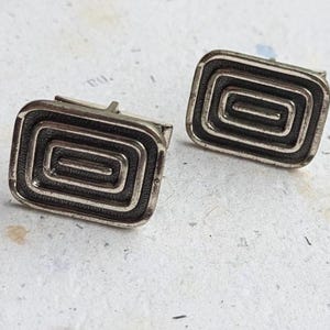 May include: A pair of silver cufflinks with a geometric design. The cufflinks are rectangular and have a concentric circle pattern.
