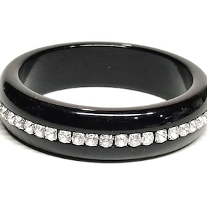 May include: A smooth, polished black band ring featuring a single horizontal row of small, clear, round-cut sparkling stones embedded around the center of the outer circumference, creating a contrast between the dark surface and the light.