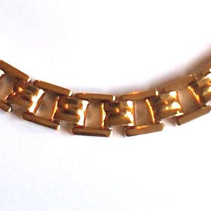 May include: A gold-toned necklace with a repeating rectangular link design. The links are connected by smaller, rounded bars, creating a geometric pattern. The necklace is set against a white background, highlighting its metallic sheen.