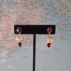 May include: A pair of vintage earrings with a gold-tone metal design. Each earring features a round, red gemstone at the top and a teardrop-shaped red gemstone at the bottom. The earrings have a leaf-like design with clear accents.