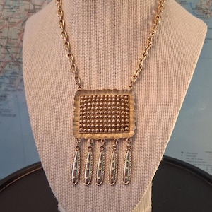 May include: A gold-tone necklace featuring a rectangular pendant with a textured, beaded surface and a scalloped edge. Five elongated, tapered drops with vertical ridges and small dot accents hang from the bottom of the pendant on a thick chain.