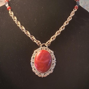 May include: A gold-toned necklace featuring a large, oval, red pendant with a decorative silver-colored frame. The chain is a textured gold color, and the necklace has small red and blue beads near the clasp. The necklace is displayed against a black background.