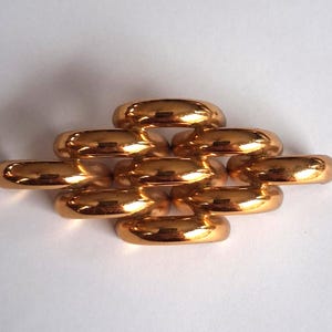 May include: A collection of ten polished, gold-colored rings arranged in a brick-like pattern. The rings have a smooth, rounded design and reflect light, creating highlights and shadows. The rings are displayed on a white surface.