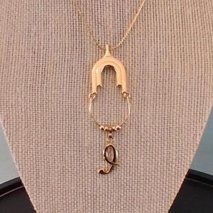 May include: Gold-toned necklace with a pendant featuring an arched design and a dangling letter "Q". The necklace is displayed on a neutral-colored surface, showcasing its delicate details and elegant style.