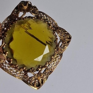 May include: Vintage gold-toned filigree ring with a large, faceted, yellow-green gemstone. The ring features intricate openwork metal detailing around the stone, creating a delicate and ornate design. The gemstone has a rectangular shape.