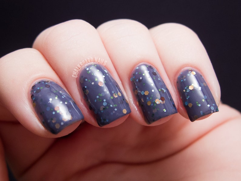 以下が含まれることがあります： ダークブルーグレーのマニキュアとイリデッセントグリッターでペイントされた、手入れの行き届いた爪のクローズアップ。爪は丸く滑らかで、背景には「CHALKBOARD NAILS」の文字が見えます。